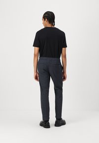 Camiseta negra de manga corta, pantalones oscuros de cuadros con cinturilla elástica y zapatos negros, vistos desde atrás contra un fondo liso.