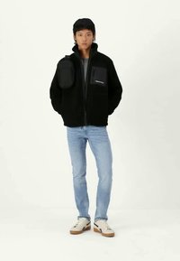 Black fleece jacket with a zip, featuring a gray patch pocket and an attached small pouch, paired with light blue jeans and white sneakers.