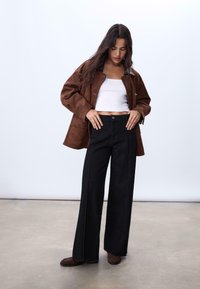 Veste oversize en daim marron, top court blanc, pantalon large taille haute noir avec des détails de couture, et mocassins marron.