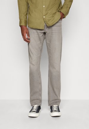 Homme portant un jean gris clair, des baskets noires et blanches, et une chemise à manches longues vert olive boutonnée, debout avec une main dans la poche.