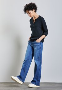 Schwarzes Langarmhemd mit Knopfleiste, kombiniert mit blauen weit geschnittenen Jeans und weißen Sneakers; steht mit den Händen in den Taschen.