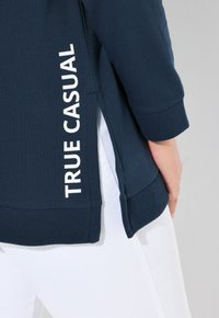 Maglietta a maniche lunghe blu scuro realizzata in tessuto a coste, con testo bianco "TRUE CASUAL" sul lato e fessure laterali per un tocco in più.