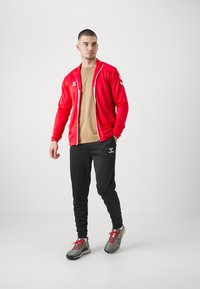 Sarkans kabatas žakets ar rāvējslēdzēju, bēša t-krekls, melnas sporta bikses un pelēki sporta apavi. Žakets ar baltajiem akcentiem; sporta biksēm logo uz augšstilba.