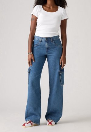 Vrouw draagt een wit kort mouwtopje, hoge taille wijde blauwe denim cargobroek, rode en witte sneakers, staat met ontspannen armen.