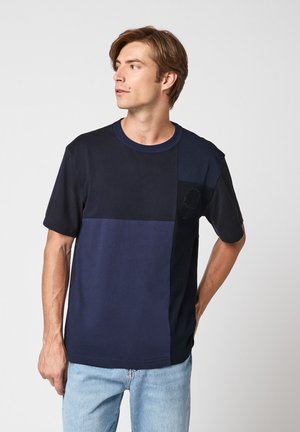 Jeune homme regardant vers la gauche, portant un t-shirt à manches courtes bleu marine avec des blocs de couleur et un jean bleu clair, sur un fond uni.