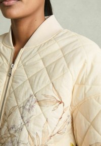 Veste bomber matelassée en tissu crème doux, avec une fermeture éclair à l'avant et des broderies florales aux couleurs douces. Col et poignets côtelés.