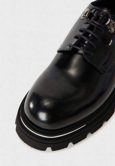 Chaussure en cuir noir avec une finition brillante, semelle noire robuste, accent blanc subtil autour de la base et système de laçage traditionnel.