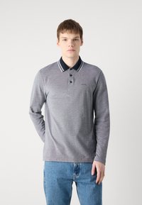 Camisa polo cinza de manga longa com colarinho azul marinho e riscas brancas, apresentando uma pala de três botões e detalhe do logótipo no peito. Usada com jeans azul claro.