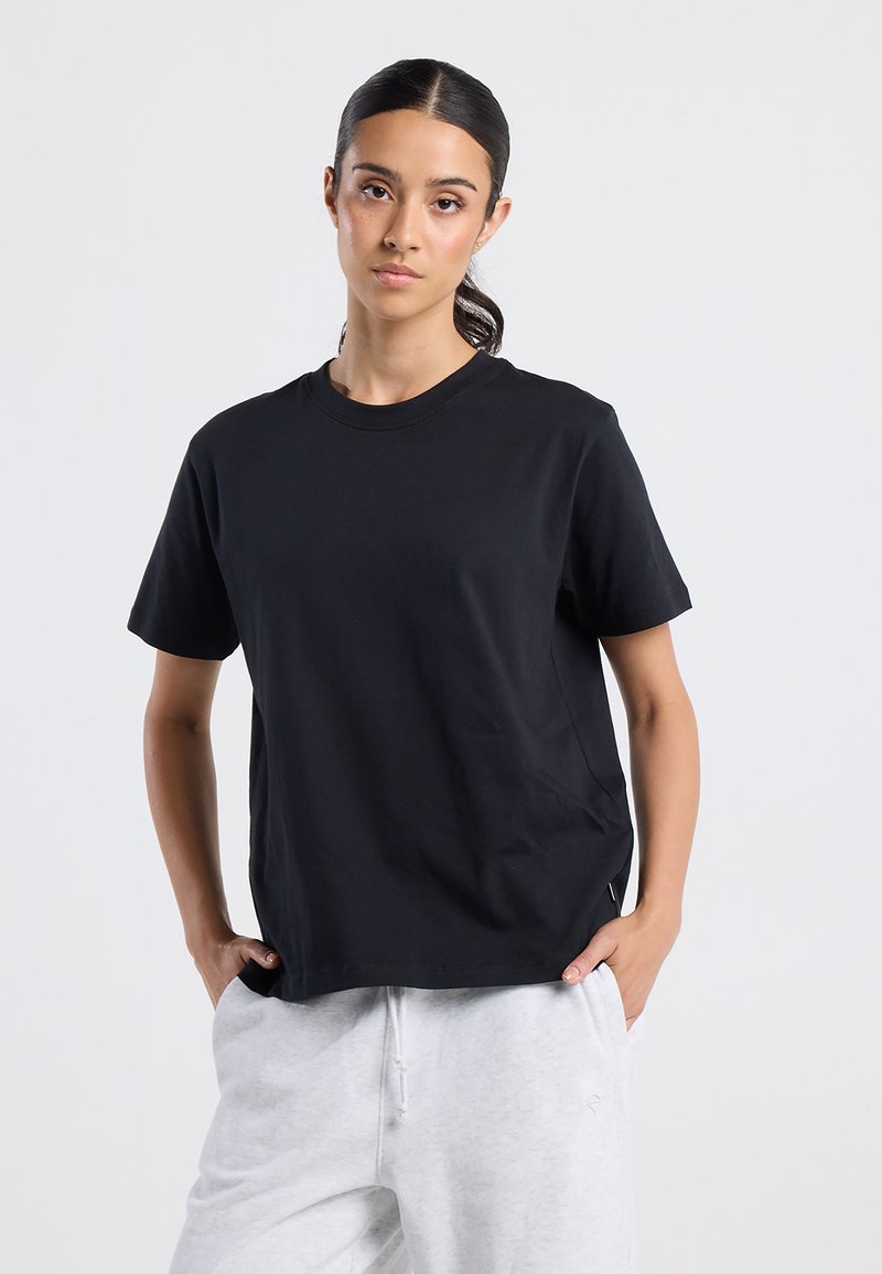 Femme aux cheveux noirs en queue de cheval portant un t-shirt noir uni à manches courtes et un pantalon de survêtement gris clair, les mains dans les poches, sur un fond neutre.