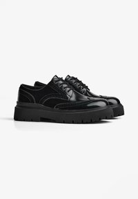 Pantofi brogue din piele neagră cu tălpi groase cu crestături, detalii tip wingtip și șireturi negre, pe un fundal alb.