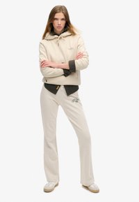 Felpa con cappuccio beige con chiusura in legno, e sottogiacca grigia; pantaloni felpati beige a trombetta con logo verde; sneakers chiare.