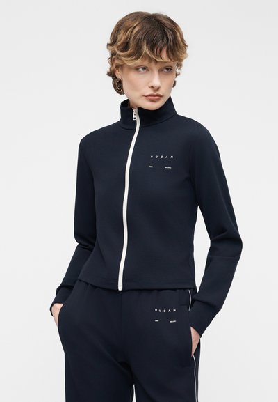 Γυναίκα με κοντά μαλλιά που φοράει μπλε σκούρα αθλητική φόρμα με λευκό φερμουάρ και το λογότυπο "Hogan" στο στήθος και στο μηρό, με τα χέρια στις τσέπες, κοιτάζοντας πλάγια.