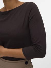 Person som har på seg en mørk brun topp med trekvart ermer, stukket inn i beige bukser med en synlig svart knapp på linningen.