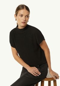 Schwarzes Kurzarmoberteil mit hohem Kragen, aus weichem Stoff mit subtilen strukturierten Akzenten. Kombiniert mit schwarzen Hosen für einen monochromen Look.