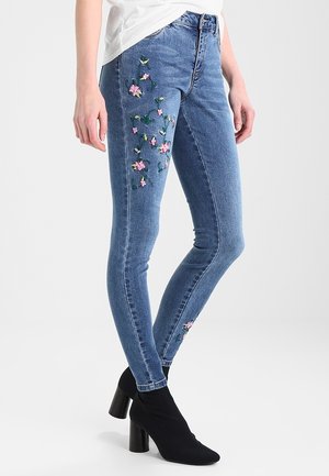 Schlanke blaue Jeans mit rosa Blumenstickerei auf den vorderen Oberschenkeln und am unteren Wadenbereich, kombiniert mit schwarzen Stiefeletten mit Absatz und einem weißen Oberteil.