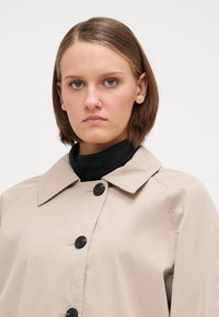 Bežový trench coat s veľkými čiernymi gombíkovými uzávermi, širokým golierom a hladkou textúrou, preložený cez vrúbkovaný čierny rolák.