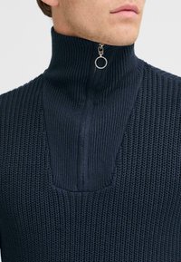 Dark navy knitted sweater with a high collar and zip closure, featuring a textured pattern and a transparent half-moon cutout.