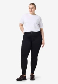 Mujer de pie con las manos detrás de la espalda, vistiendo una camiseta blanca, jeans negros ajustados y zapatos de cordones marrón oscuro frente a un fondo liso.