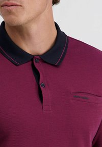 Burgunderfarbenes Polohemd mit strukturiertem Muster, dunkelblauem Kragen, zwei Knöpfen und kleinem Logo auf der Brusttasche.