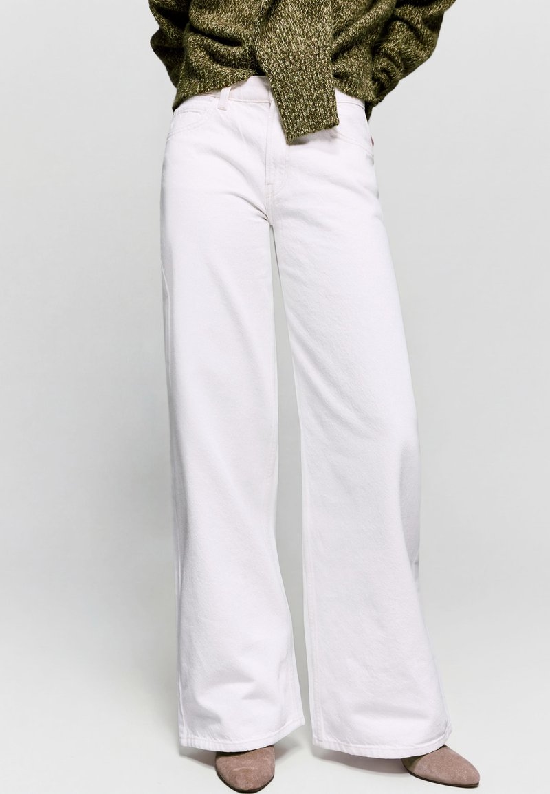 Person iført hvide vida bukser med høj talje, olivengrøn striktrøje og beige spidse sko, der står mod en ensfarvet baggrund.