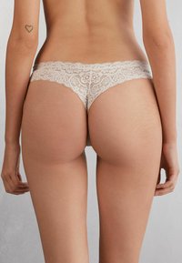 Lace string in lichtbeige, met een golvende rand en een bloemenpatroon. Gladde stoftextuur, ontworpen voor minimale bedekking.