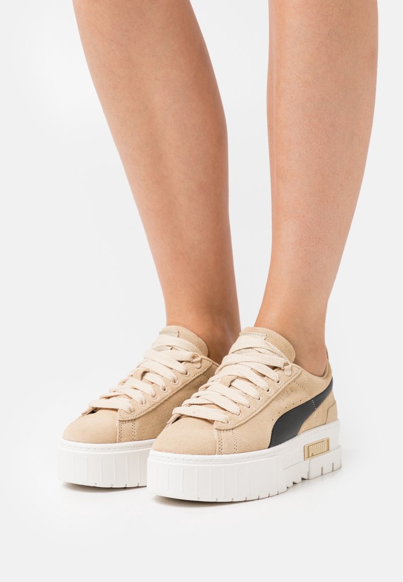 Personne portant des baskets compensées en daim beige avec des semelles blanches épaisses et une bande latérale noire, debout sur fond blanc.