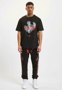 Čierne tričko s grafickým potlačou s textom "Thug Life" v červenej a šedej farbe. Čierne tepláky s červeným textom "Life" a akcentovými líniami. Biely tenisky.