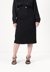 Svart denim midi-skjørt med en tettsittende silhuett, frontsømmar og en enkelt gullknapp i livet, kombinert med svarte sandaler med remmer.