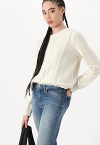 Krém kötött pulóver texturált kábelmintával, kerek nyakkal és bordázott mandzsettával, kék skinny farmernadrággal párosítva. A modell fekete dzsekit tart.