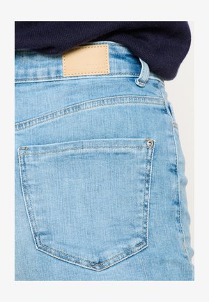 Vaaleansiniset farkut, joissa on kolmionmuotoinen takatasku, kangasvyötäröllä nahkalogo ja hienovaraisia ompeluyksityiskohtia.