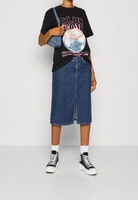 Černé grafické tričko s růžovým a bílým potiskem, spárované s modrou denimovou midi sukní a černými vysokými teniskami. Malá modrá kabelka nošená křížem přes tělo.