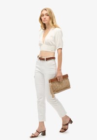 Weiße, kurz geschnittene Bluse mit V-Ausschnitt und Spitze, kombiniert mit engen weißen Jeans, einem braunen geflochtenen Gürtel und einer gewebten Clutch. Braune Sandalen mit Absatz.