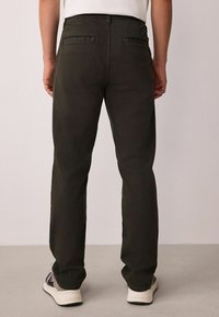 Dark green trousers with a straight leg design, featuring two back pockets and visible stitching details. Worn with white and grey sneakers.