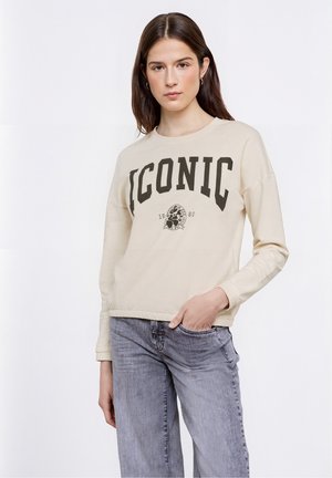 Felpa color crema con la scritta audace "ICONIC" in grigio scuro, con dettaglio grafico e fondo a costine. Abbinata a jeans grigi scoloriti.