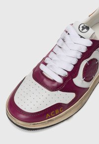 Sneaker in pelle bordeaux e bianca con punta forata, lacci bianchi, patch circolare con logo e suola in gomma testurizzata. Marchio dorato “ACBC” visibile.