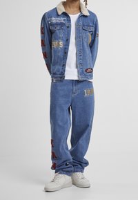 Denim jacket with a shearling collar, blue color, patches, and embroidered text. Paired with matching pants featuring red accents and numbers.