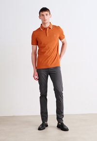 Oranžinė polo marškinė su baltais akcentais, derinami su pilkomis siaurėjančiomis kelnėmis ir juodais basučių tipo batais. Slydus paviršius, kasdieniška forma, minimalus prekės ženklo ženklinimas.