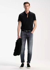 Camisa polo negra con acentos en el collar marrón, combinada con jeans gris desgastados. El modelo sostiene una chaqueta negra y lleva zapatillas negras. Diseño simple.
