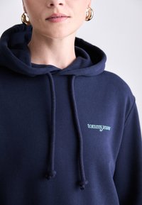 Haut à capuche bleu marine en tissu doux, avec une poche kangourou, des cordons de serrage et le logo "tommyjeans" en broderie bleu clair sur la poitrine.