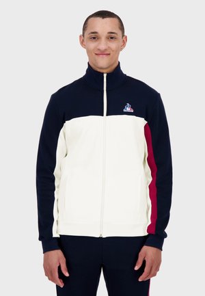 Navy and cream zip-up jacket with high collar, featuring maroon side stripes and a logo on the left chest. Soft fabric and fitted design.