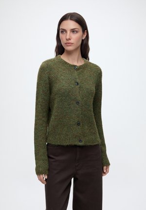 Femme portant un cardigan en laine vert boutonné et un pantalon marron foncé, debout devant un fond clair uni.