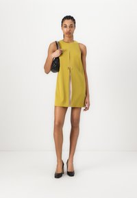 Yellow sleeveless dress with a center slit, styled with a black shoulder bag and black high heels. Smooth texture and minimalistic design.