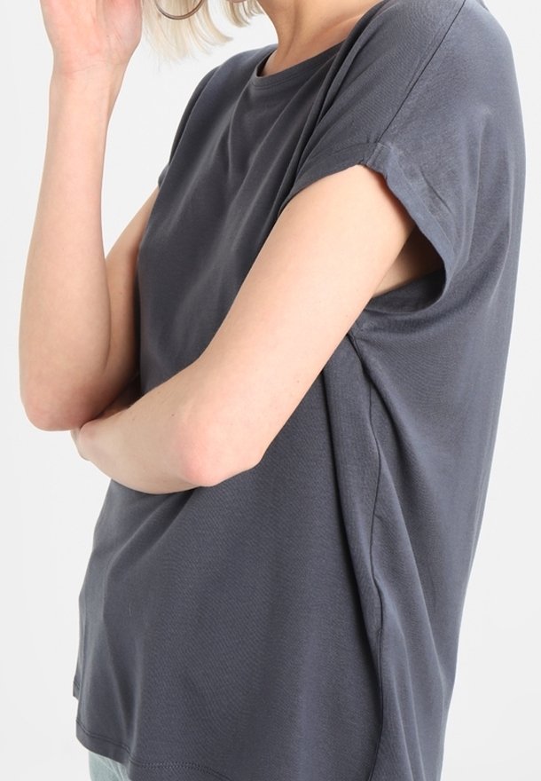 Personne portant une chemise ample gris foncé à manches courtes, les bras croisés et une main touchant son visage.