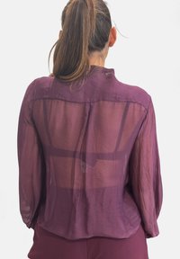 Blusa viola trasparente con vestibilità rilassata, maniche lunghe e pannello posteriore con motivo a griglia. Tessuto leggero e dalla texture liscia.