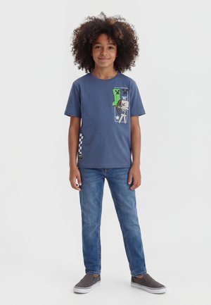 Barn med krøllet hår iført blå Minecraft grafik t-shirt, blå jeans og grå slip-on sko står på hvid baggrund.