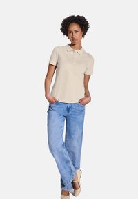 Beige pikétröja med krage och treknapps-stängning, kombinerad med ljusblå vida jeans. Klädseln har en avslappnad och ledig passform.