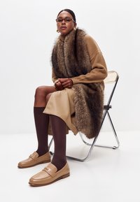 B�éžový pletený svetr, hnědý umělý kožich, béžová kožená sukně, hnědé punčocháče a béžové mokasíny s logem, sedící na kovové židli.