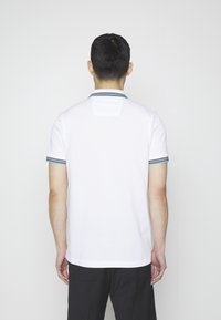 Homme debout de dos portant un polo blanc à manches courtes, avec un col et des bords de manches rayés, et un pantalon noir.