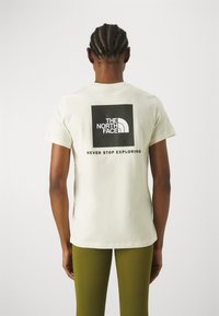 Osoba s copy v vlasech, oblečená v bílém tričku s logem "The North Face" a nápisem "Nikdy nepřestávejte objevovat", v kombinaci s olivově zelenými kalhotami.