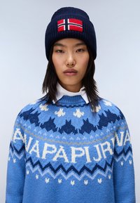 Maglione azzurro lavorato a maglia con motivi geometrici bianchi e blu scuro; berretto blu scuro con un design della bandiera norvegese rosso, bianco e blu.